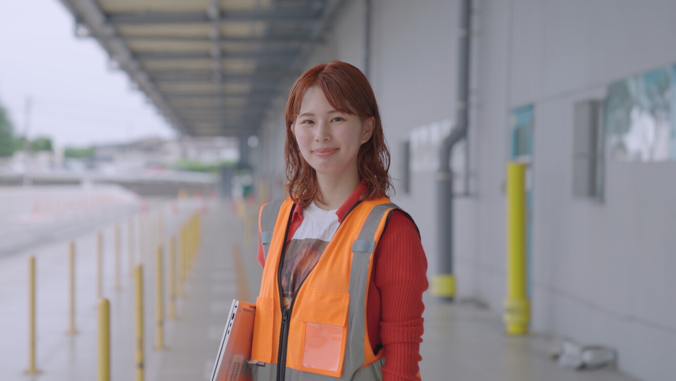 オレンジ色の安全ベストを着ている明るい茶色の髪の女性（三木悠さん）が配送センターの屋外に立っている。