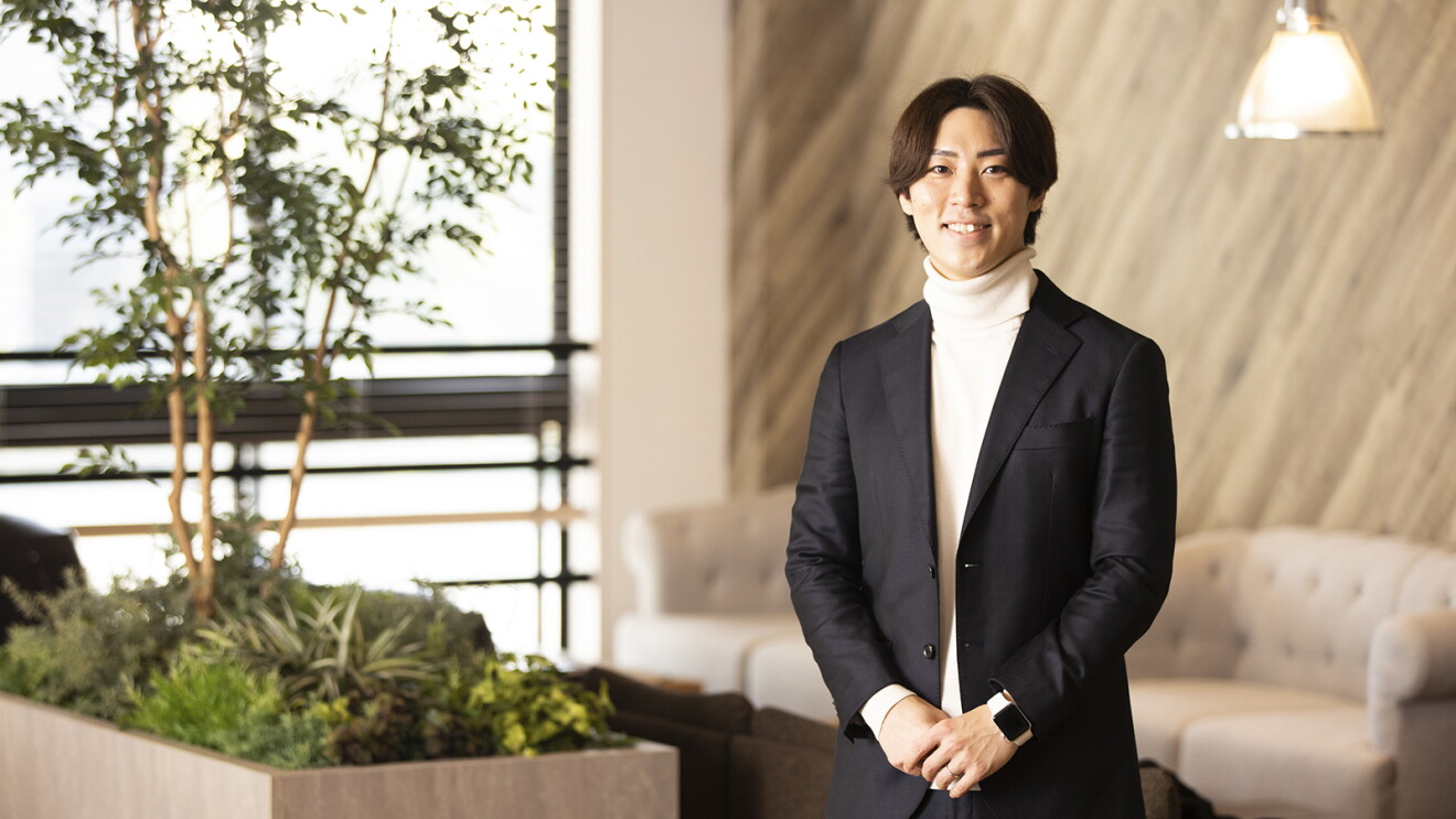 木材を基調とした落ち着きのあるインテリアのオフィスを背景に笑顔で立つジャケットを着た杉谷さん