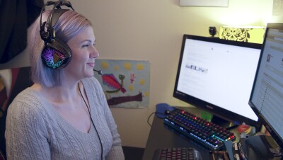 A woman wears a headset while looking at two computer monitors.