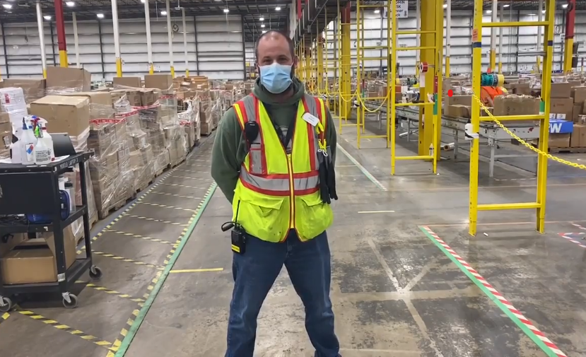 Russel Whitfield, an associate at Amazon, stands in an Amazon warehouse smiling for a photo. He is wearing a face mask and other Amazon warehouse safety gear. 