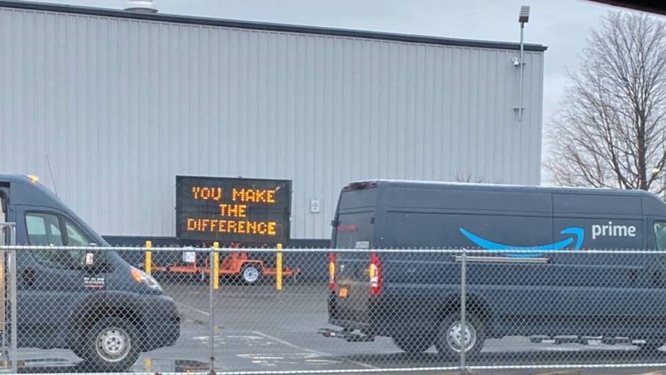 Samochody dostawcze Amazon Prime na parkingu centrum realizacji. W tle znajduje się znak drogowy z napisem „Ty robisz różnicę”.