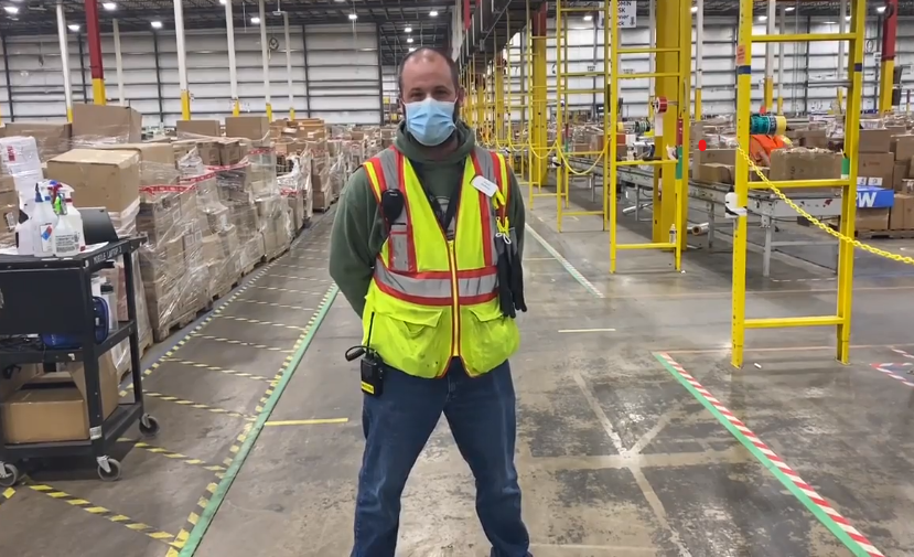 Russel Whitfield, an associate at Amazon, stands in an Amazon warehouse smiling for a photo. He is wearing a face mask and other Amazon warehouse safety gear.