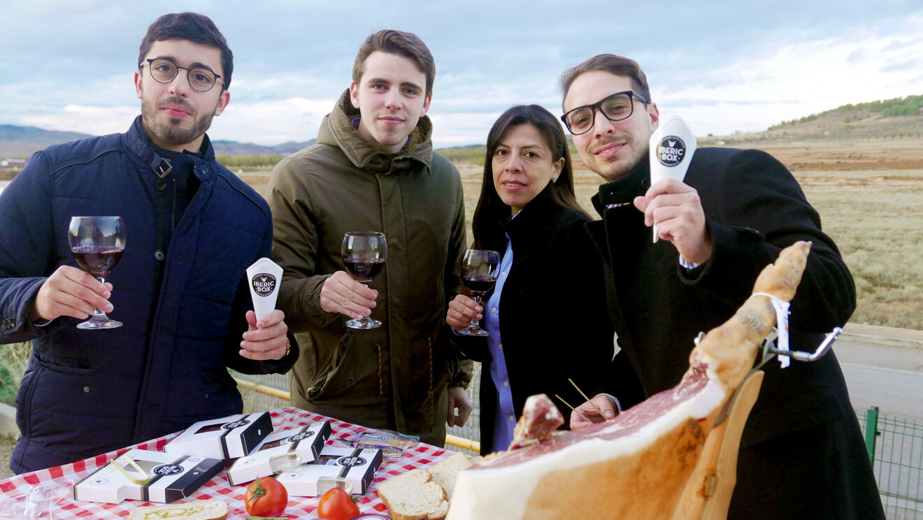 El equipo de Iberic Box, 4 jóvenes que no superan los 40 años, están posando detrás de un mesa lleva de comida y brindan con copas de vino tinto. En la izquierda, el CEO de la startup, David González, con una copa de vino tinto y un sobre de jamón, acompañado de tres colegas del equipo. Todos visten con chaquetas de invierno. La mesa tiene un mantel de cuadros, un jamón con el jamonero, un plato de jamón, las cajas de Iberic Box, una botella de aceite y otra de vino. El fondo de la foto es el cielo azul. 