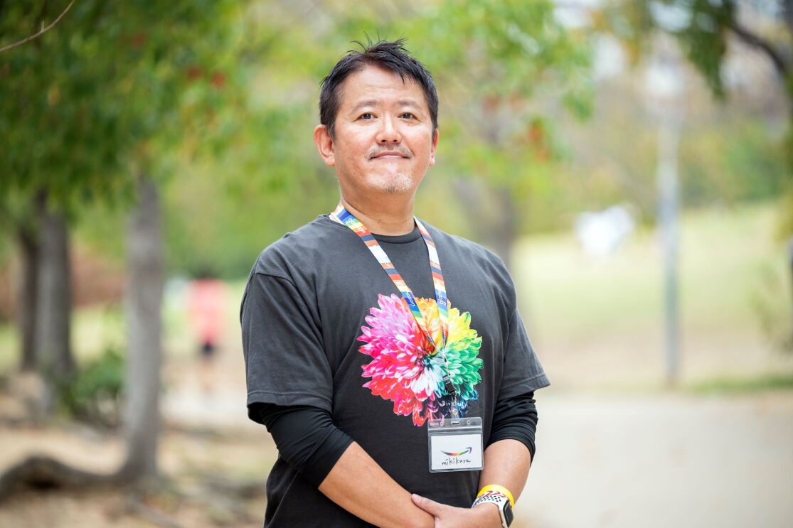 カラフルな花が描かれている黒いTシャツを着ている男性(倉橋充樹さん)が手を組んで、左側に少し傾いて立っている。