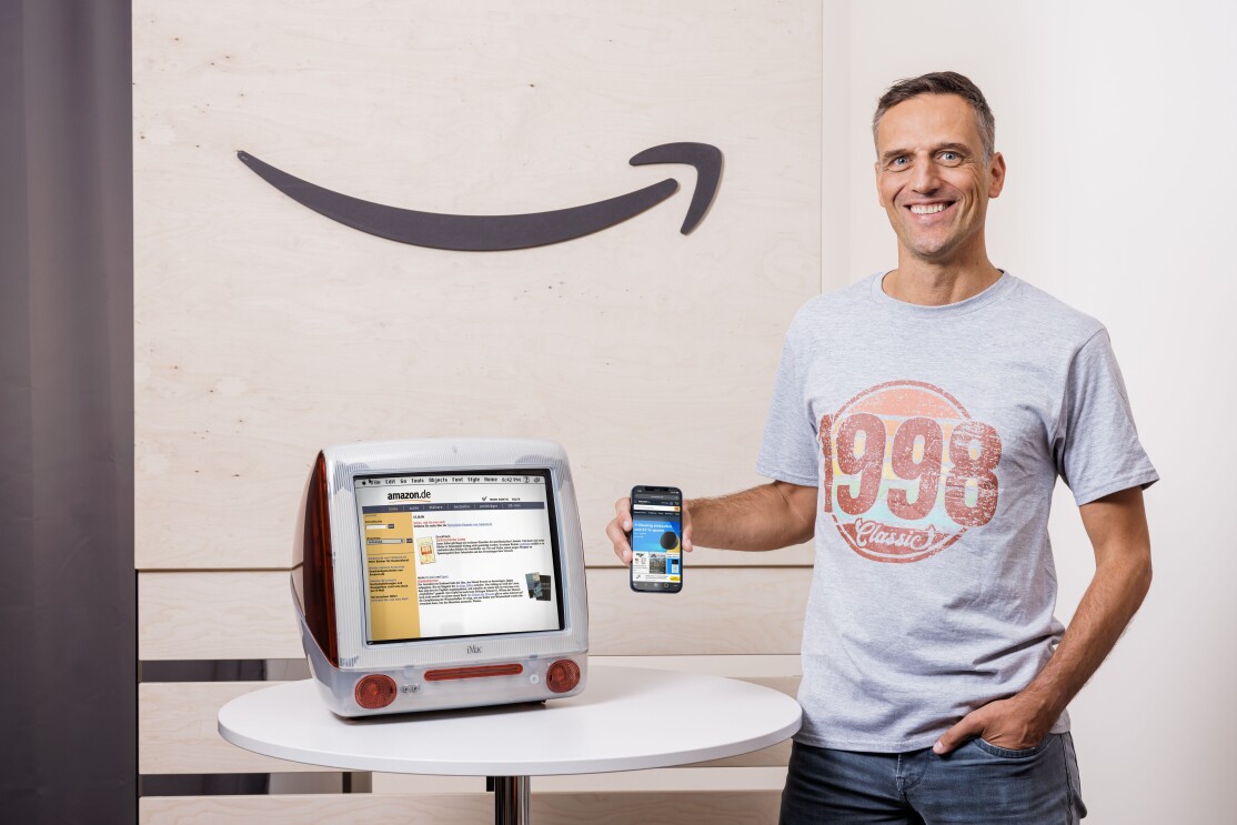 Amazon Deutschland gibt es seit 25 Jahren. Rocco Bräuniger hält ein Smartphone mit der Amazon App vor einen PC mit der Amazon Website.