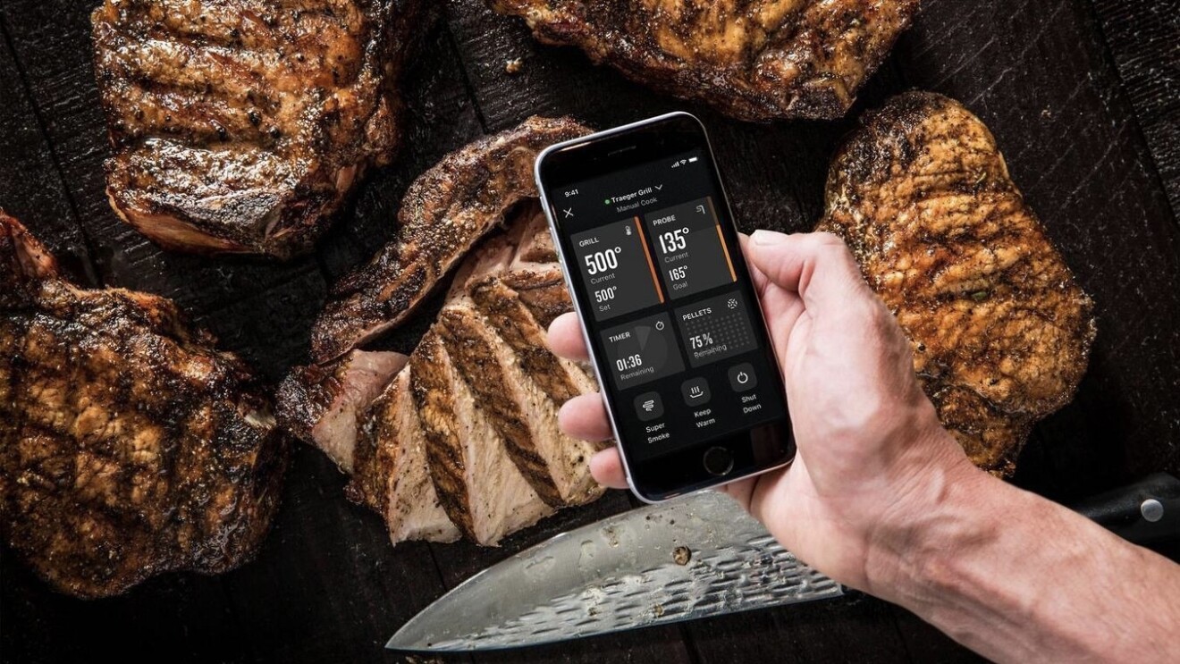 An image of a man holding a phone with the Traeger app open. Behind the phone in the photo is five large, grilled steaks and a large knife that has cut into one of the steaks. 