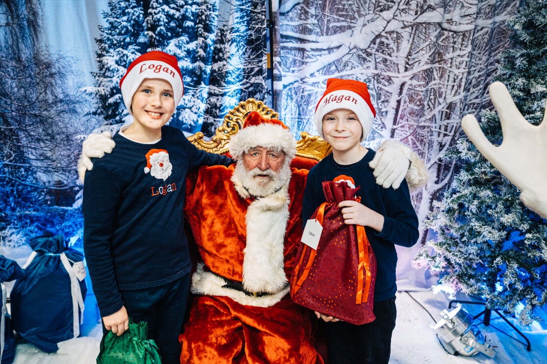 Two Children standing either side of Santa Claus in Santa's Grotto. They are each wearing Christmas hats. 