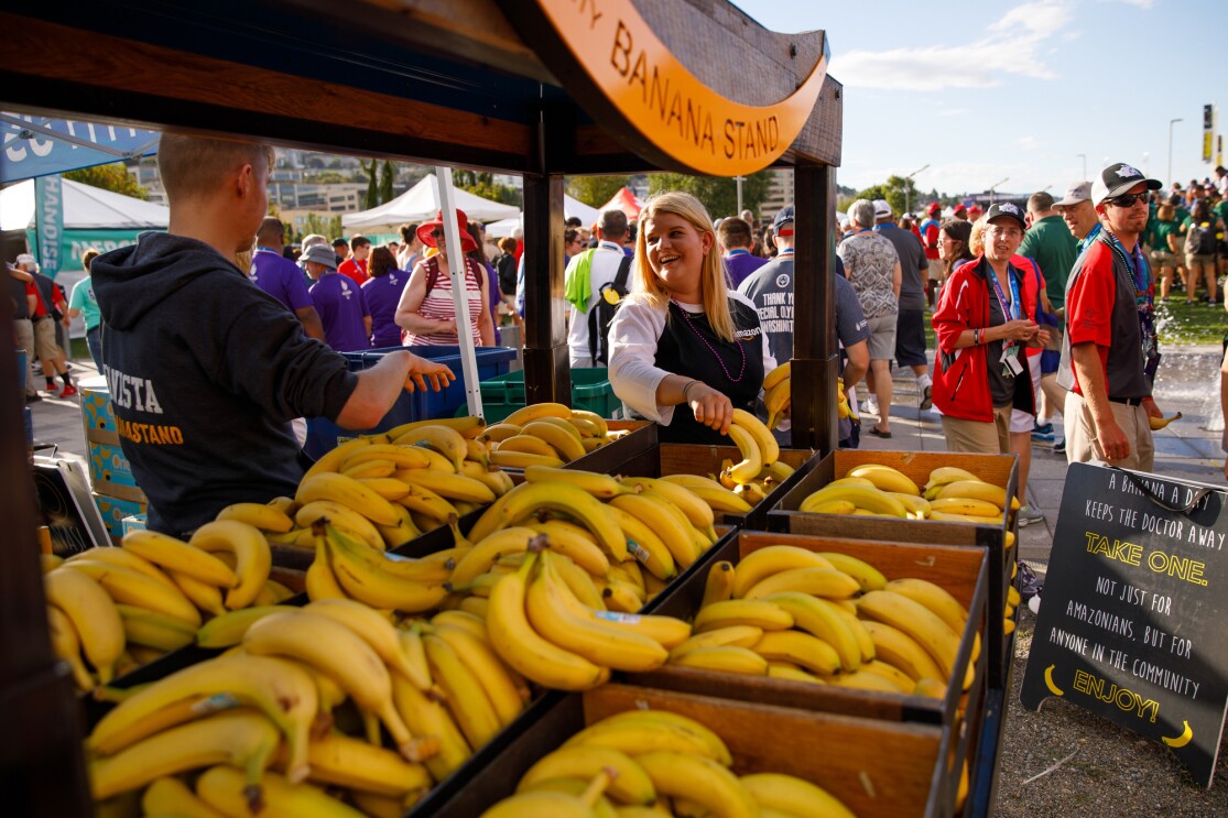 The Community Banana Stand is shown in the foreground of this photo, with stacks of bananas available for attendees, for free, attended by two "bananistas." In the background, event attendees are seen walking and standing.