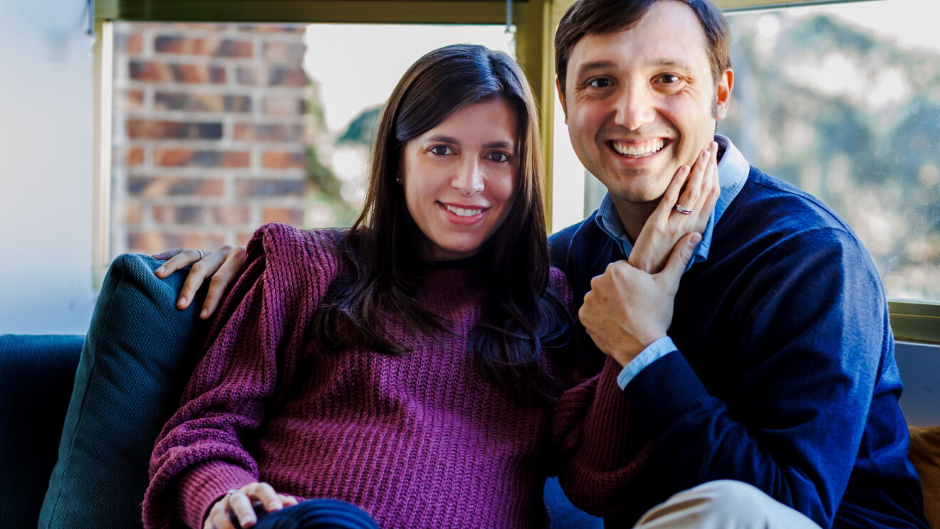 Miriam y Alberto sentados en el sofá de su casa. El sofá es de color gris y estña delante de un ventanal. Ella va con un jersey de punto morado, el pelo largo castaño hasta el pecho y tiene su mano izquierda apoyada sobre la cara de Alberto. Se le nota la barriga de embarazada. La otra mano la está apoyando su pierna. Él tiene el brazo en la espalda de Mirian. y agarra la mano que su mujer le tiene en la mejilla. Alberto lleva un pantalón de color beige, un jersey azul oscuro y una camisa azul clarito. Los dos están mirando a cámara y sonriendo. 