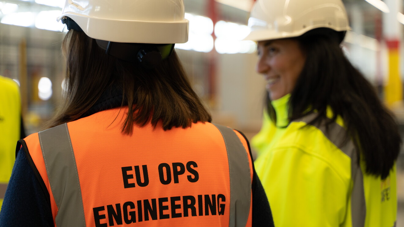 Sonia Corvera, Responsable Senior de Construcción para Amazon España, e Isabel Bermúdez, Responsable Senior de Proyectos de Ingeniería para Amazon España. Las dos protagonistas están de espaldas y llevan casco. Sonia va con un chaleco amarillo e Isabel naranja. Se están mirando y Sonia está sonriendo.