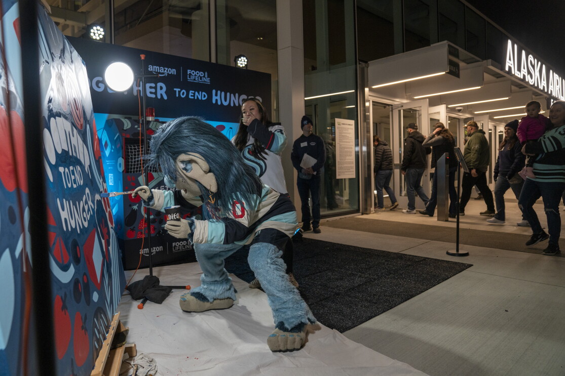 Seattle Kraken mascot, Buoy, takes a turn painting on artist Stevie Shao's mural outside of the Climate Pledge Arena, as Stevie watches from behind.