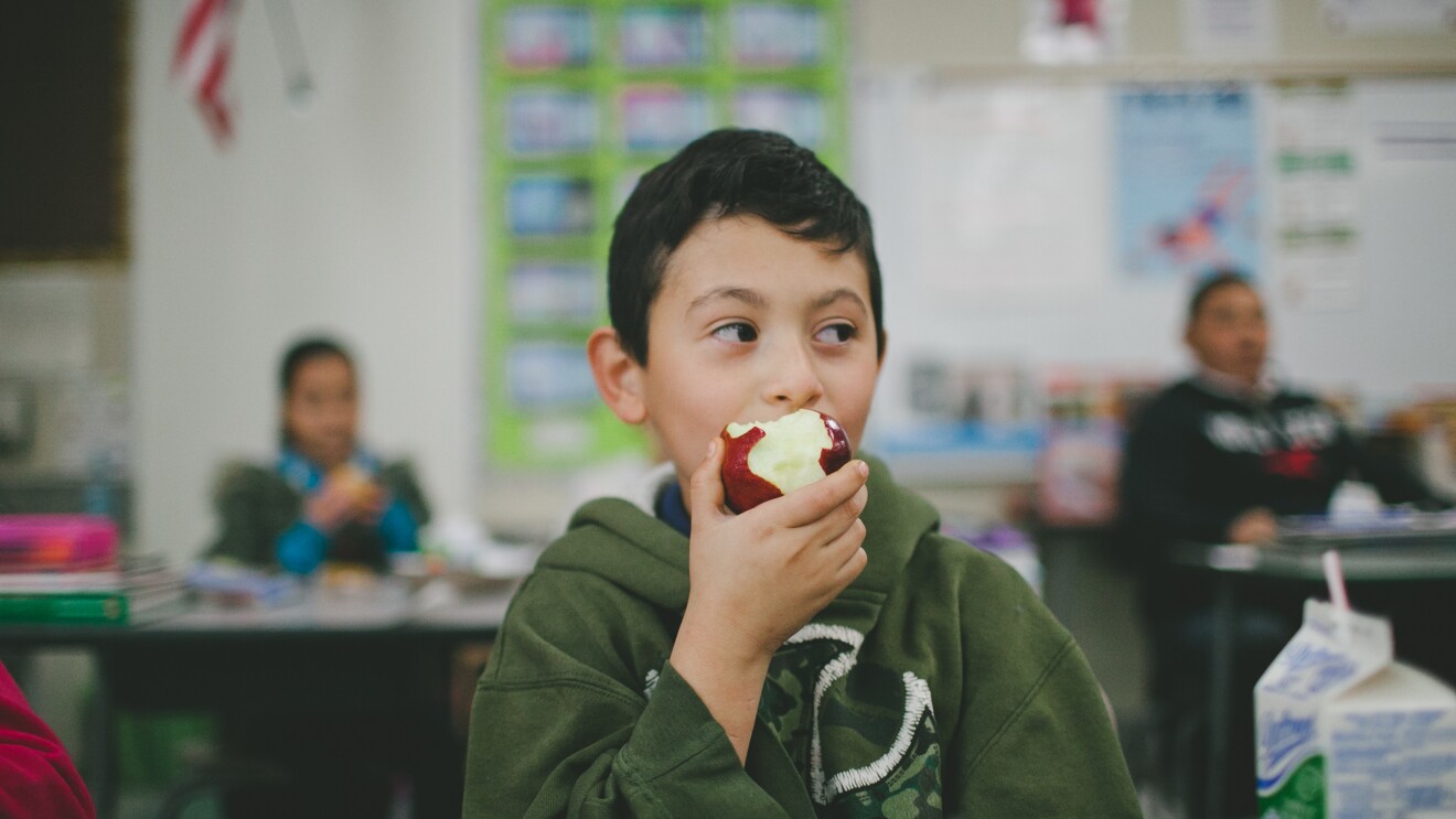 一个男孩在教室里吃苹果。在他身后,其他学生都坐在桌子。