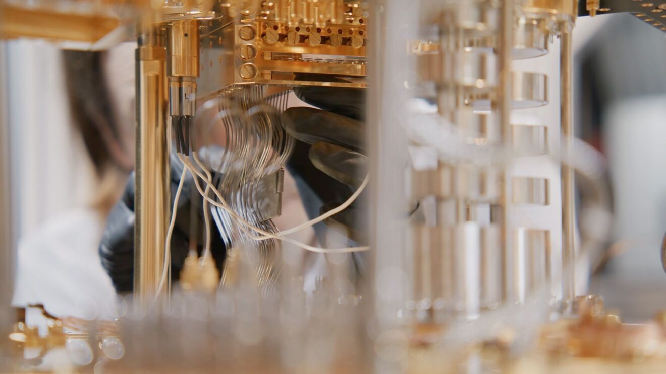 金色の金属部分やガラス管、ワイヤーが複雑に組み合わされた実験装置。装置の奥側には作業者がいて、黒い手袋をつけた手で装置に触れている。