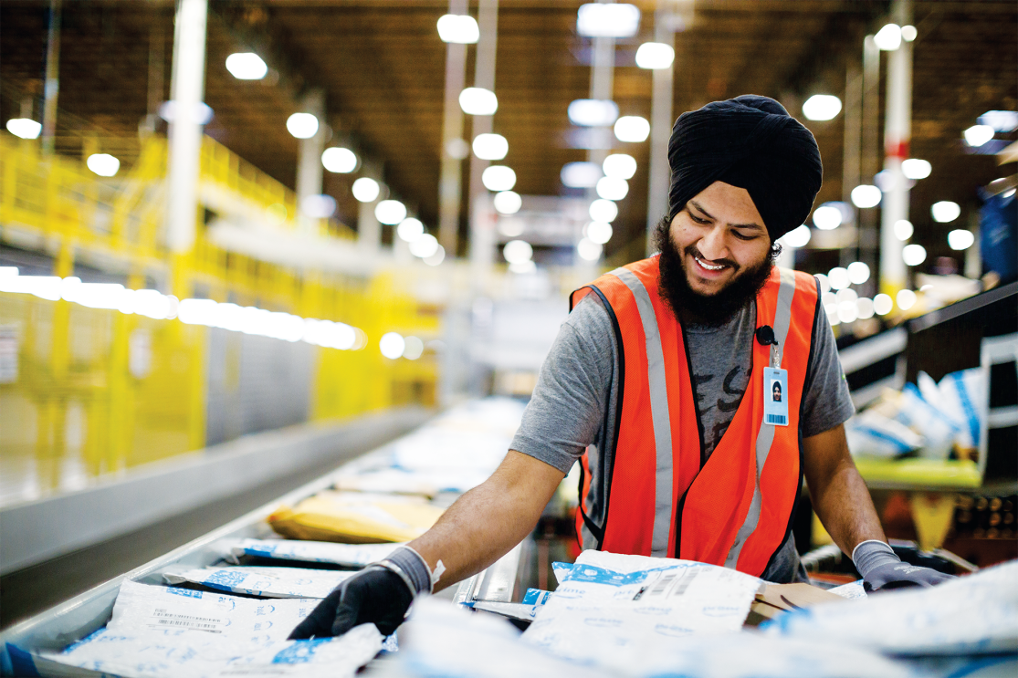A man, wearing a dastaar (or turban) in an Amazon fulfilment center (warehouse). He is moving packages from a conveyor belt to a large cart to be shipped.