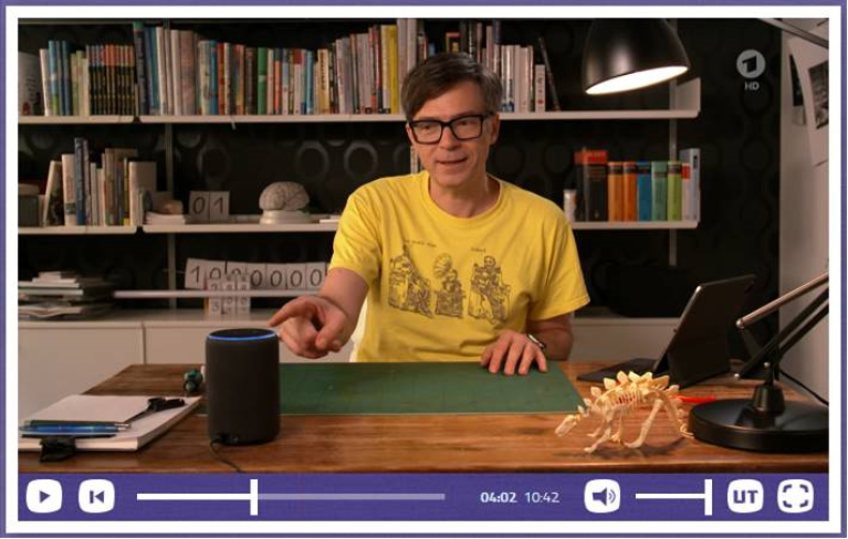 Ein Mann in einem gelben Tshirt sitzt an einem Schreibtisch und zeigt mit dem Finger auf  ein Alexa Echo.