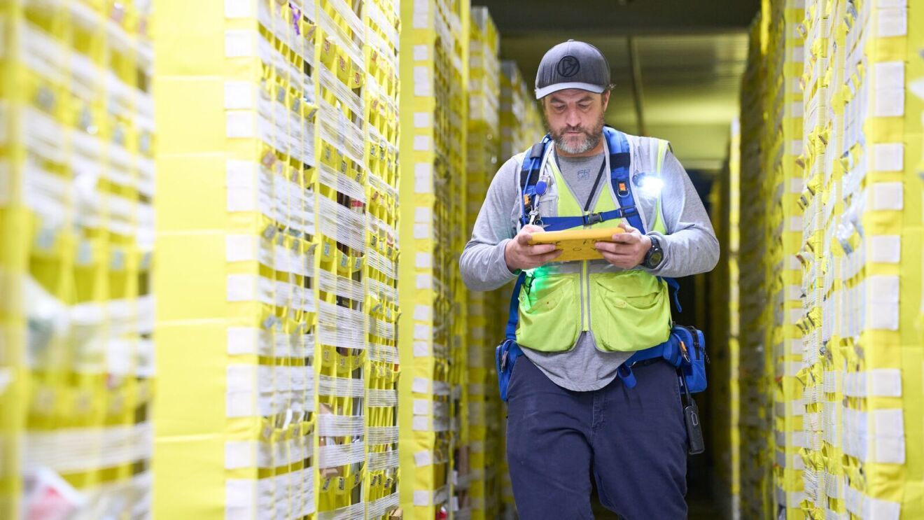 man wearing a cap and safety vest walking through shelves of products and inspecting a list inside an amazon fulfillment center