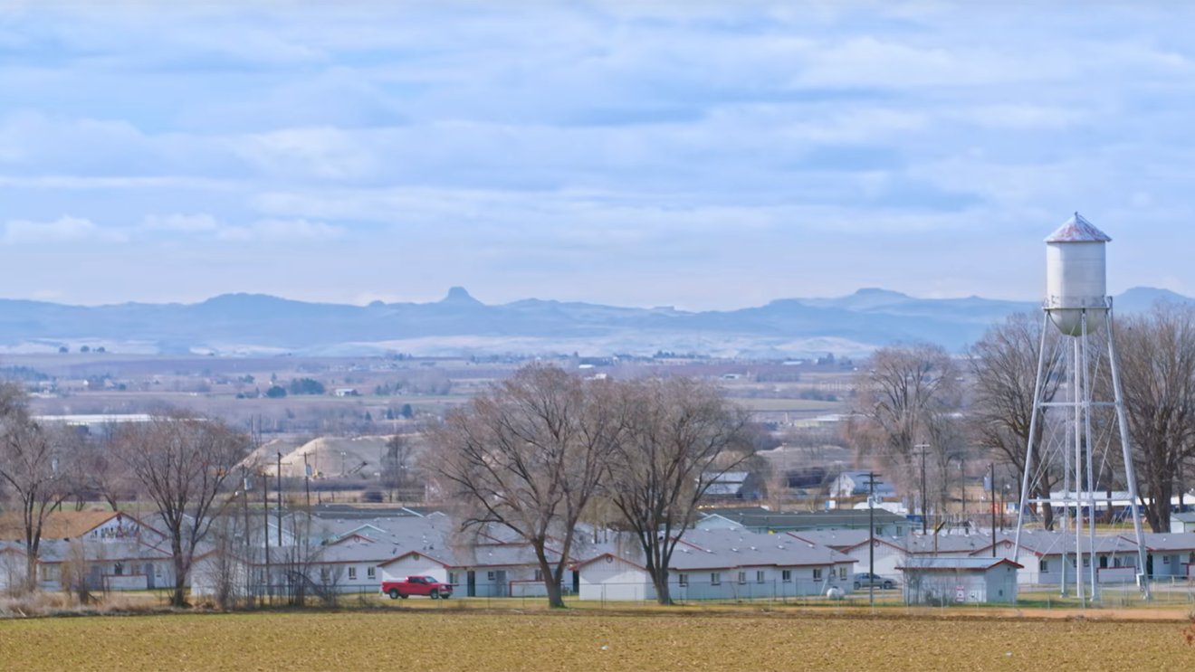 A picturesque view of a rural Idaho town. In the foreground, small trees dot the landscape, and a water tower juts into the sky, near several small ranch homes. In the background, foothills rise up behind the landscape.