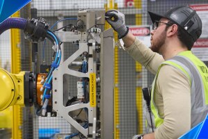 An Amazon employee wears a yellow safety vest and works on a robot.
