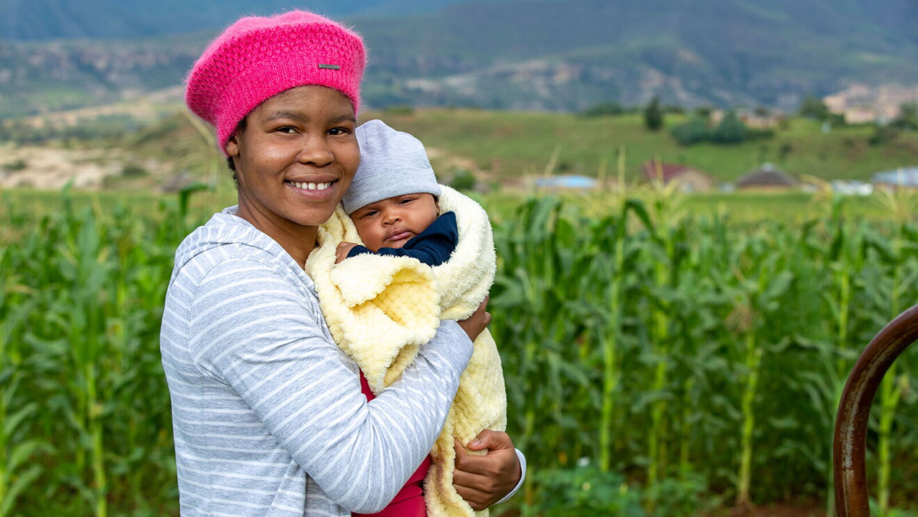 An image of a woman smiling for a photo while standing in front of a green field. She is holding a newborn baby wrapped in a light yellow blanket.