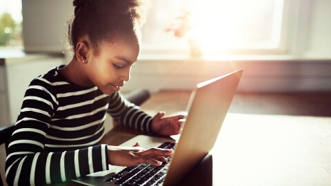 A young girl sits at a table typing on her laptop.