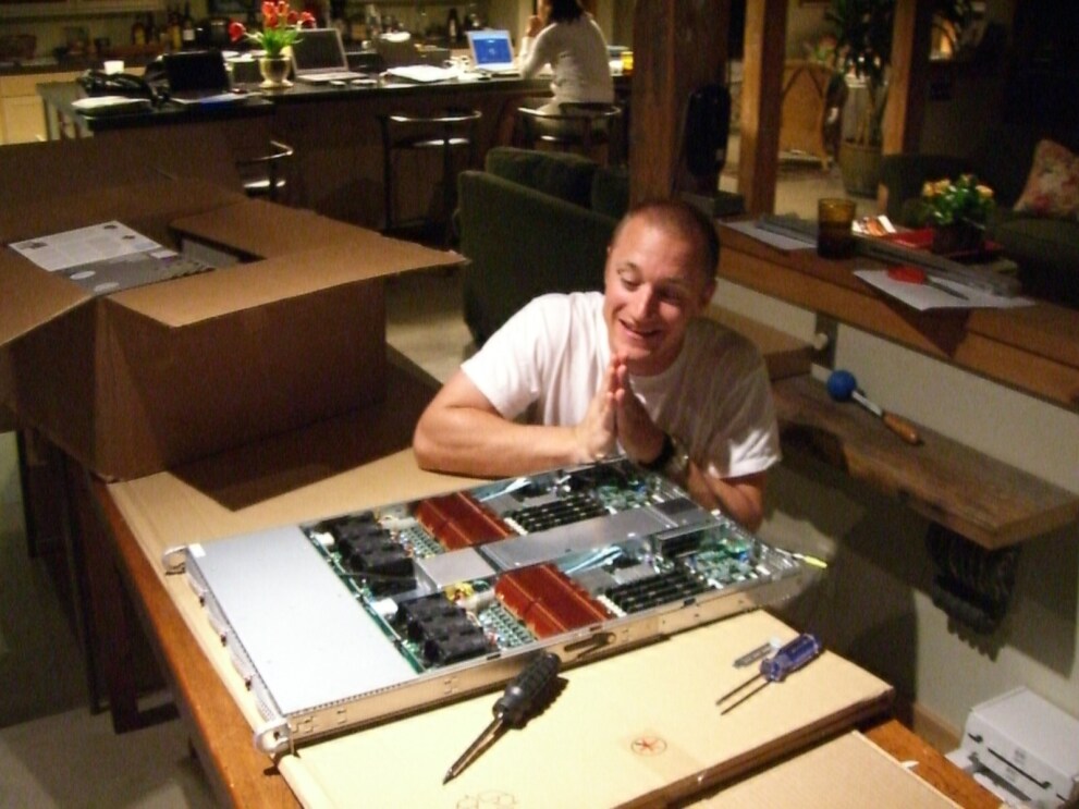 An image of a man holding his hands in a prayer position while sitting next to a server he just built.