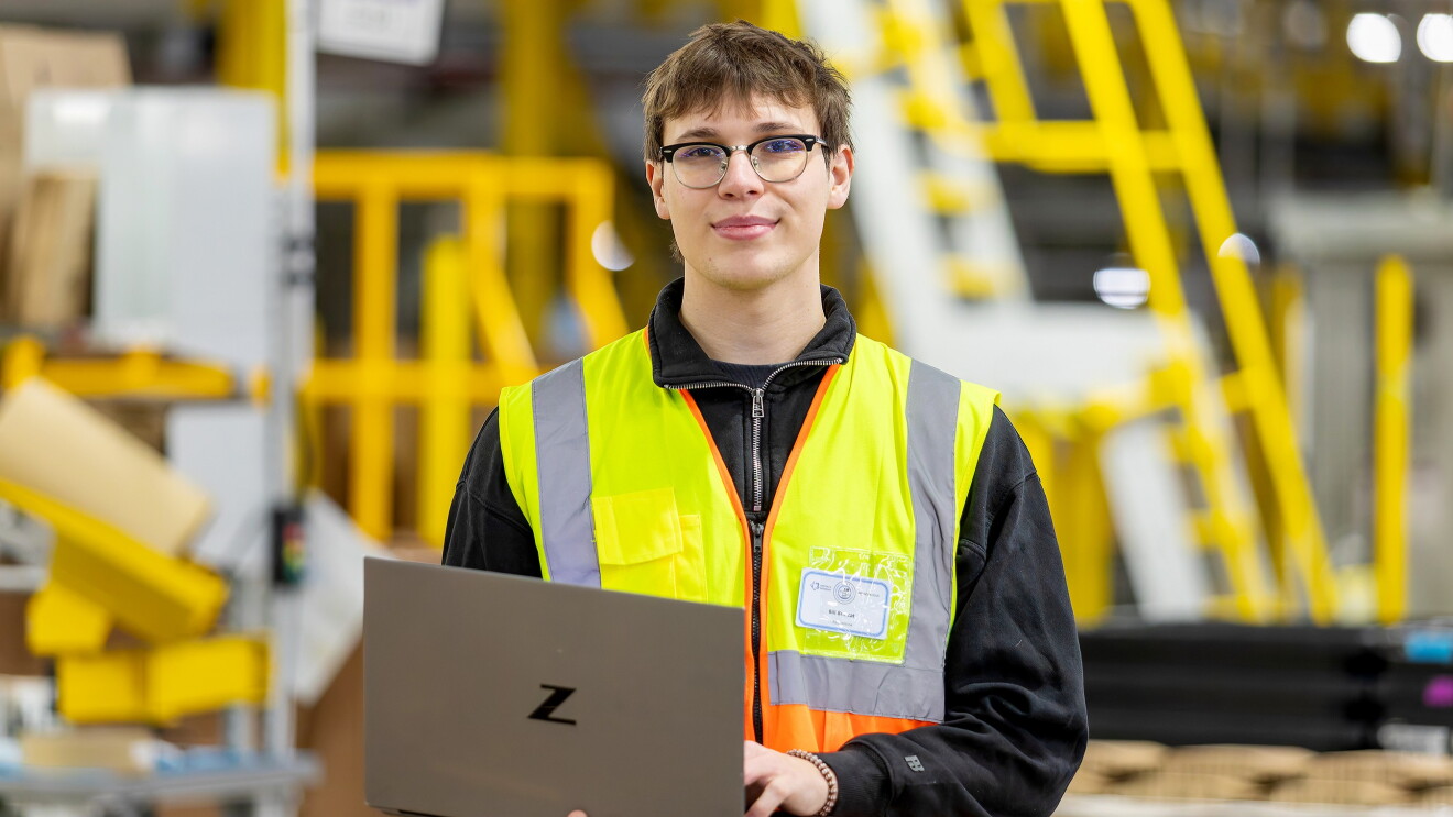 Young man wearing high vis and holding a laptop in a warehouse background