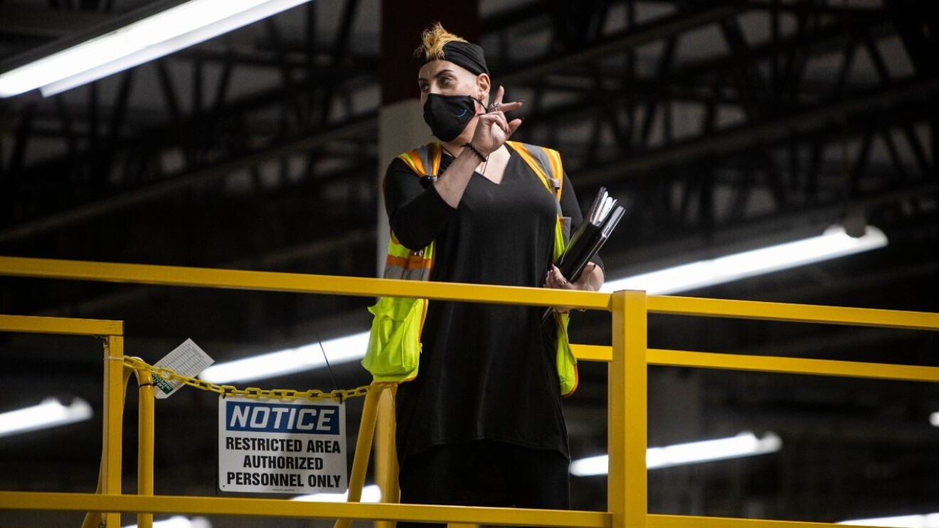 An image of an amazon employee standing in a fulfillment center wearing a mask and a safety vest while giving instructions to fellow employees.