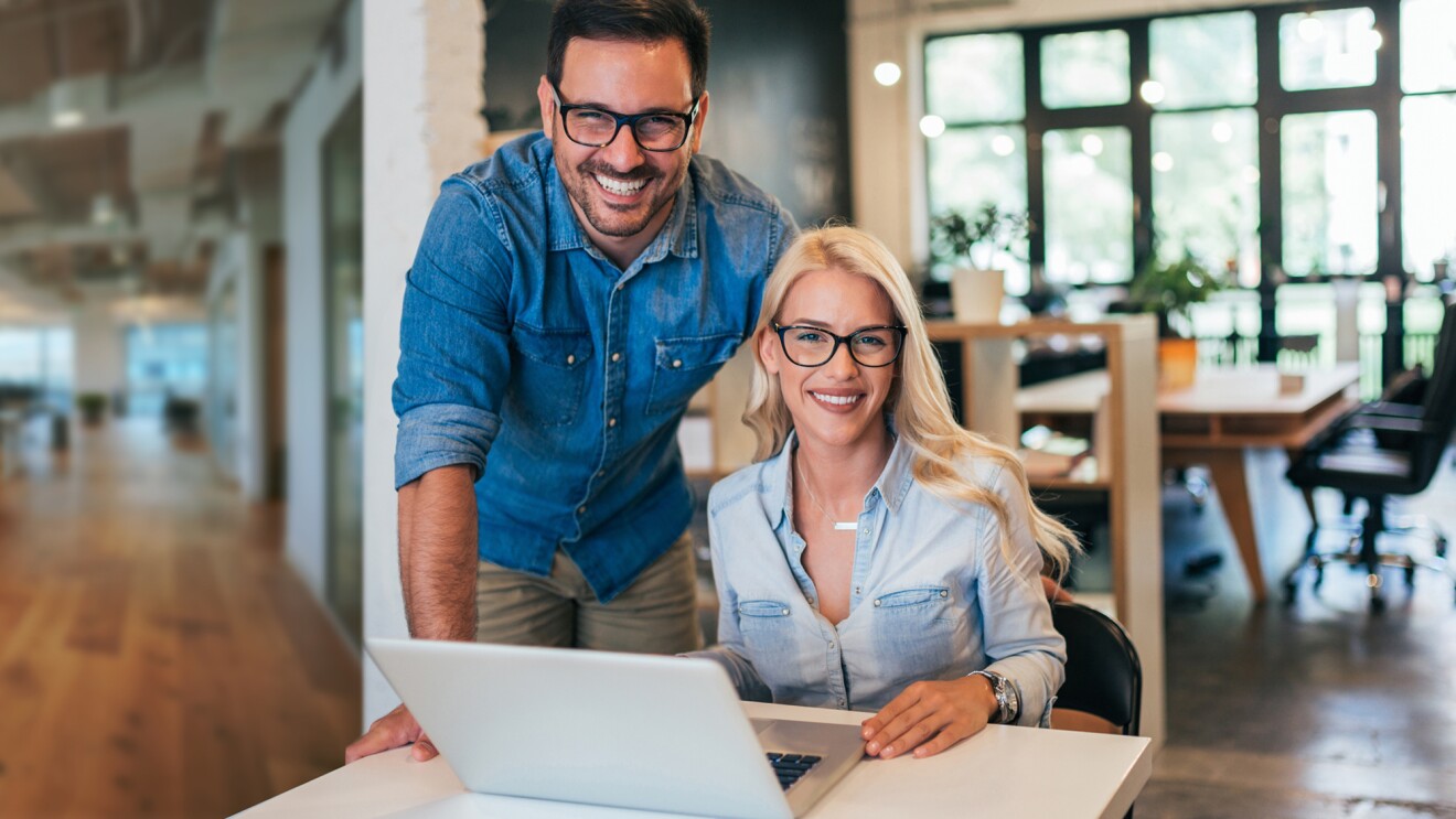 A photo of two business owners. One is sitting at a desk, that has a laptop device on it. The other is standing behind the person who is sitting down.