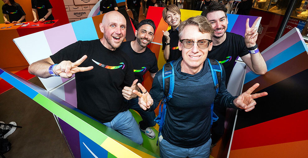 5人の人物が大きな箱の中に入って笑顔でポーズを取っている。5人のうち4人はAmazonロゴ入りのお揃いのTシャツ。大きな箱はレインボーカラーやトランスカラーに塗られている。
