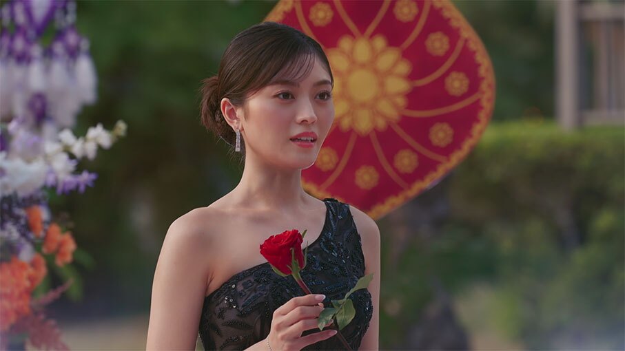 生花や装飾的な布が飾られた屋外に立つ女性。ワンショルダーのドレスを着用、体の前で赤いバラを持っている。