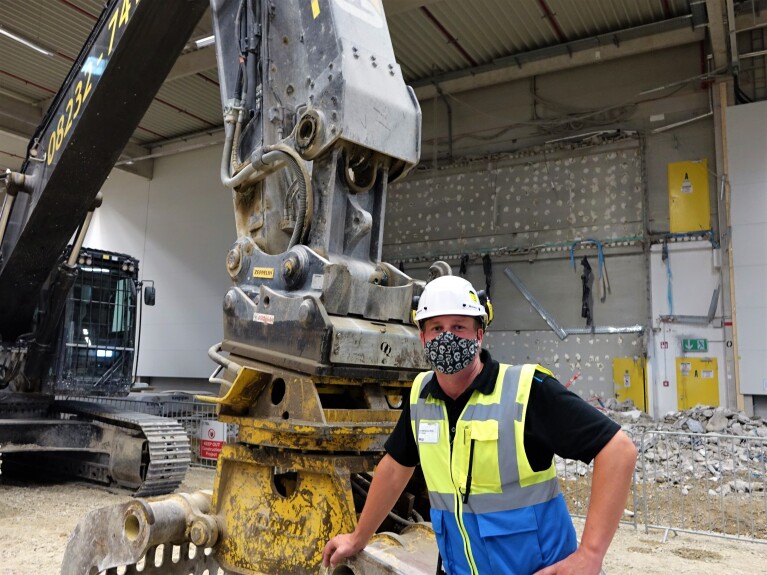 Oliver Reisenhofer mit Maske und Helm neben einem Bagger auf der Baustelle
