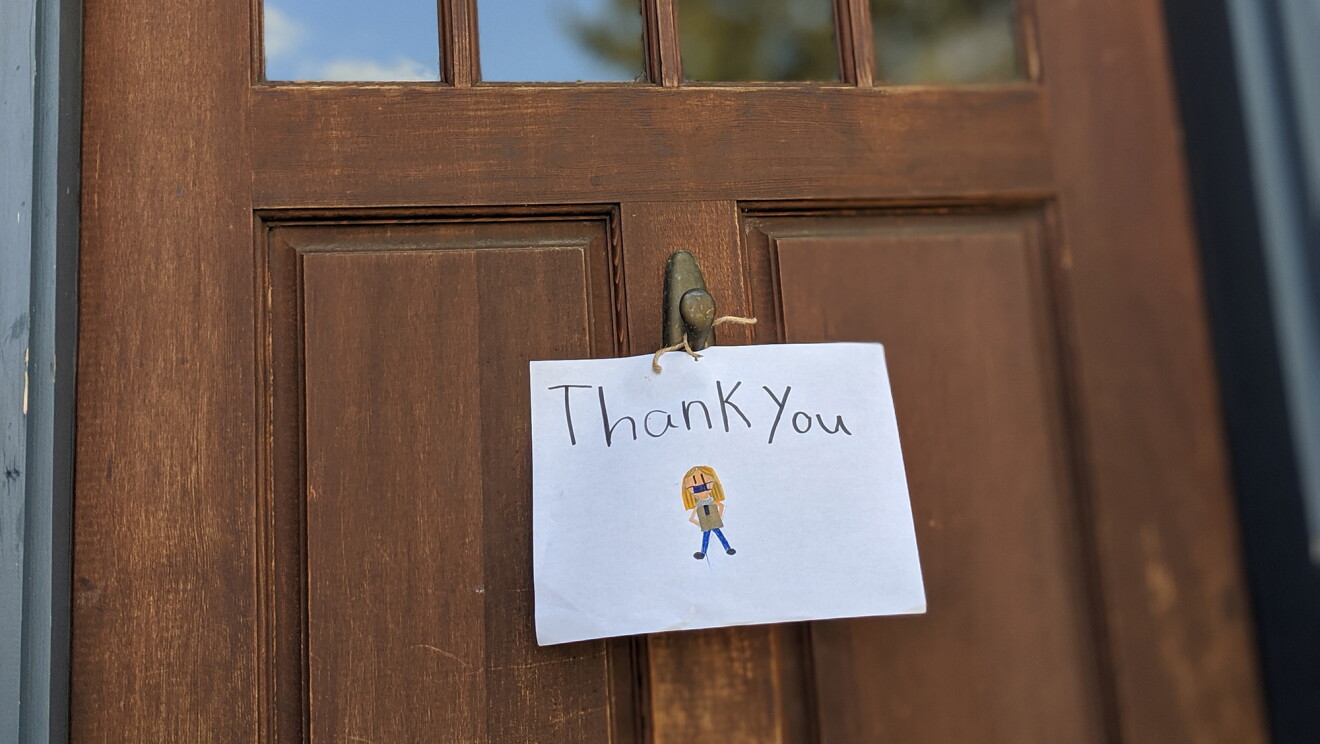 A sign attached to the front door of a home, with a delivery driver wearing a mask, and "Thank you" drawn on it. 