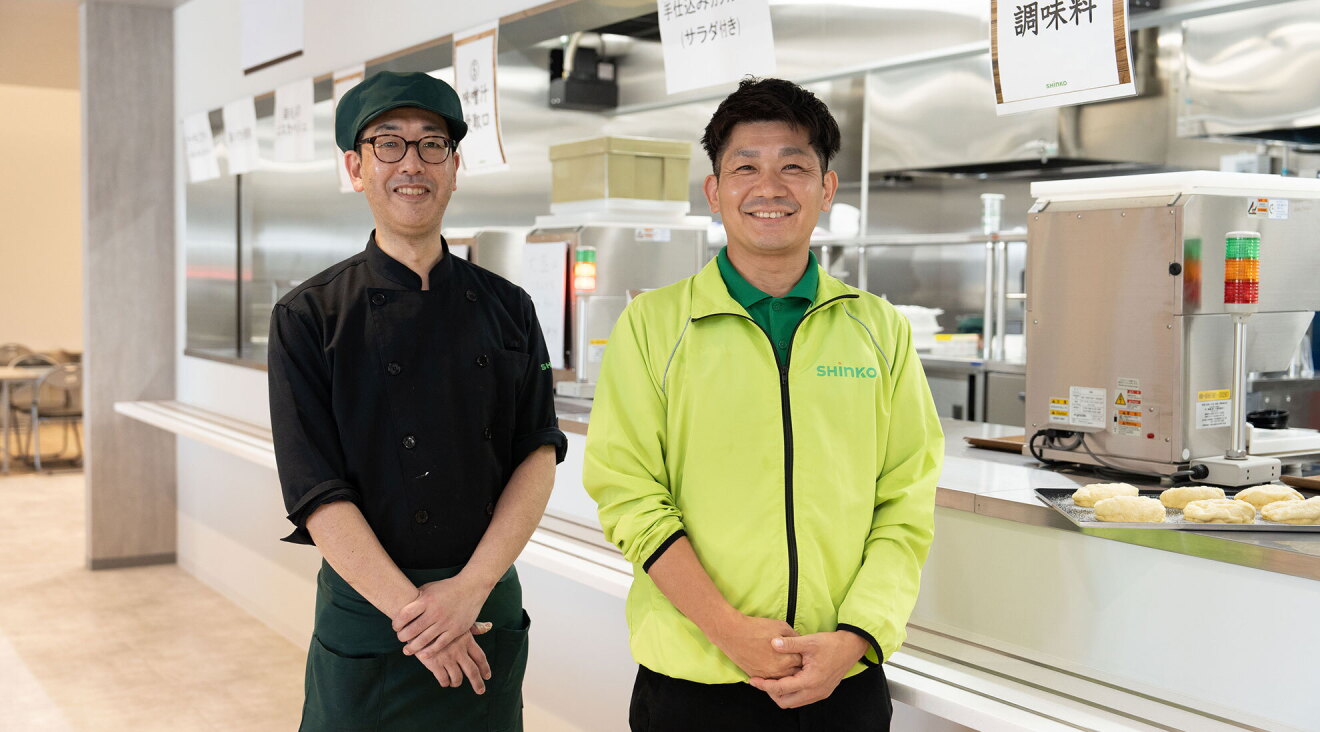 食堂の前に立つ2人の男性一人は黒い調理用服を着ている