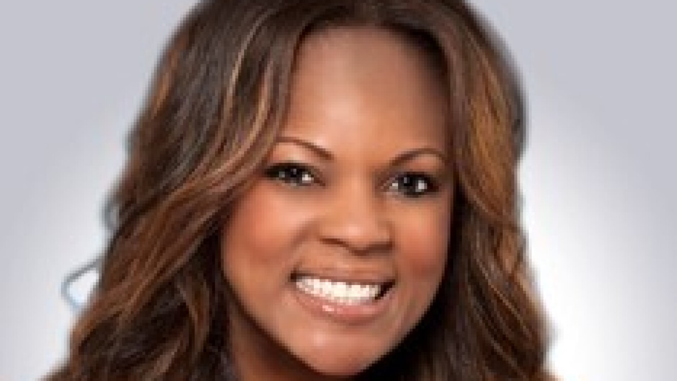 A woman who is Black smiles at the camera in front of a gray screen. 