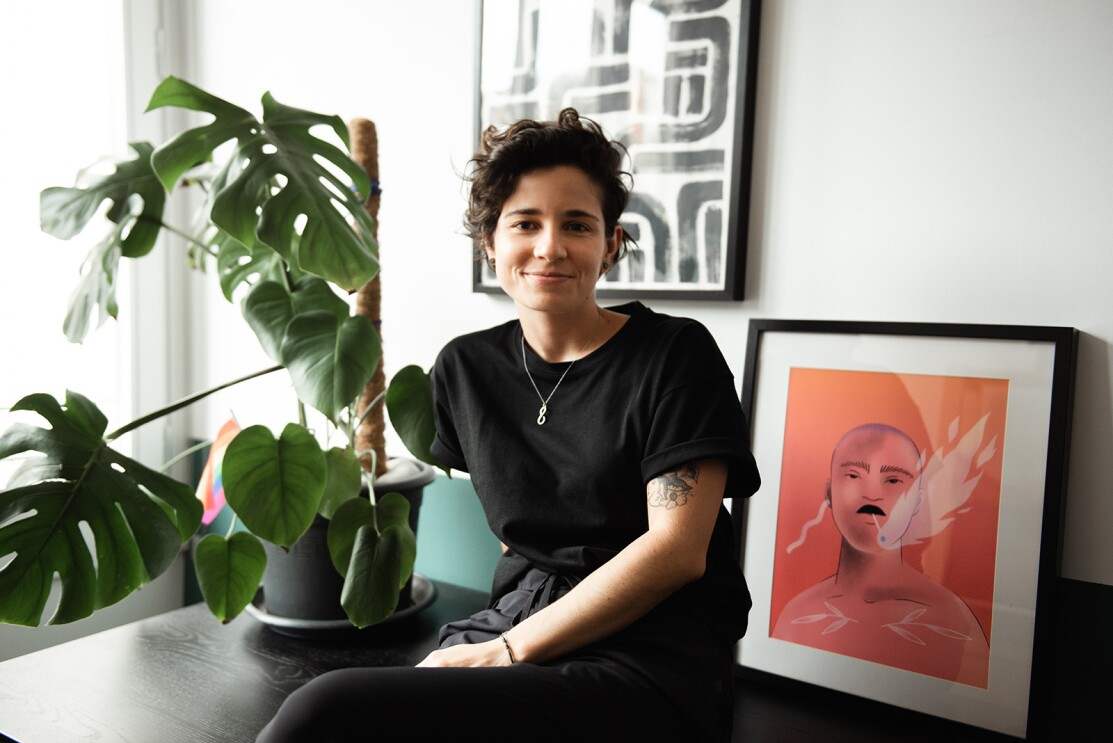 Imagem de Carmela Caldart sorrindo para a foto em uma sala de estar. Há uma planta e uma ilustração emoldurada atrás dela