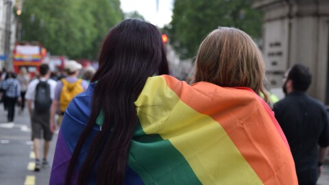 Es una foto con dos personas de pelo largo, moreno y rubio, de espaldas y tapados con una bandera del orgullo con el arcoiris. De fondo y desenfocados se ven personas andado en lo que parece un desfile. Se aprecia un autobús tojo de dos pisos con la bandera del orgullo. Por el autobús de dos pisos pensamos que es Londres.