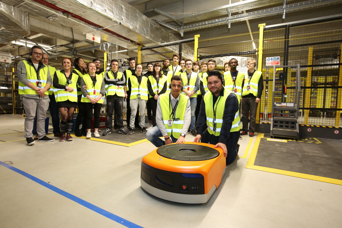 Eine Gruppe von Schülern, ausgestattet mit gelben Besucherwesten, posiert für das Gruppenfoto im Logistikzentrum. Vor ihnen: einer der Transportroboter, die in Winsen eingesetzt werden.