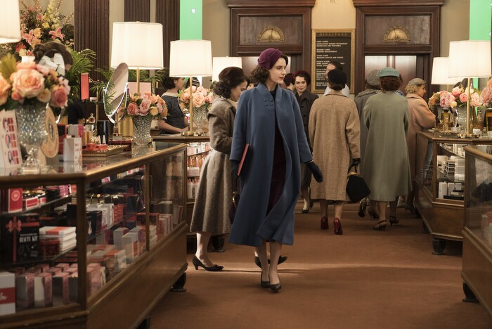 An image of Mrs. Maisel walking through a department store in a blue coat and a maroon hat. The coat is flowing as she walks.