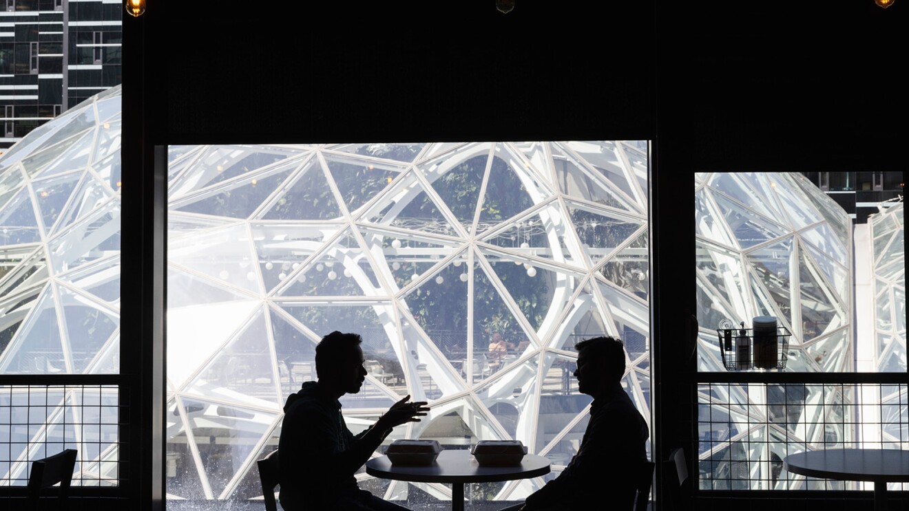 Two men are sitting at a table across from each other, talking and eating lunch. They are in silhouette, and the Seattle Spheres building looms large in the background