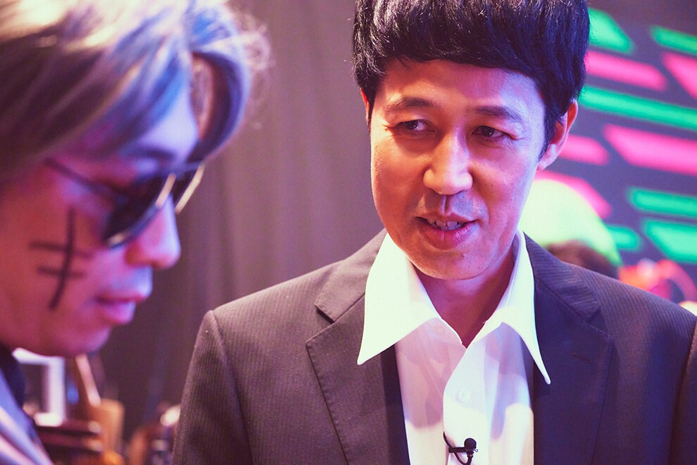 2人の男性のクローズアップ。横顔だけが写る左側の男性はサングラスをかけ、右ほほにマジックで描いた傷跡。右側の男性は黒いスーツと白いシャツを着用。背景は黒布と派手な壁。