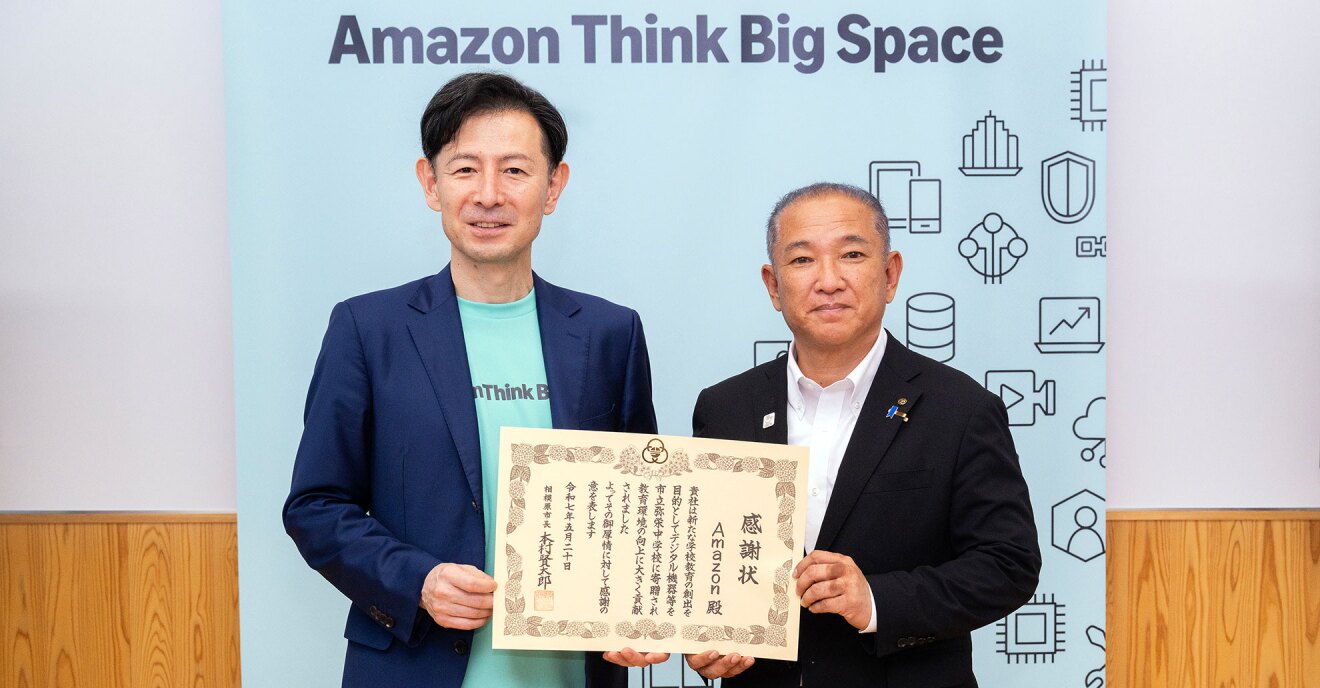 「Amazon Think Big Space」と書かれた水色の背景を前に立つ2人の人物が、1枚の賞状を手に持って見せている。左側の人物はミントグリーンのTシャツに紺のジャケットを羽織り、右側の人物はダークグレーのスーツと白いシャツを着用。賞状には「感謝状Amazon殿」と書かれている。