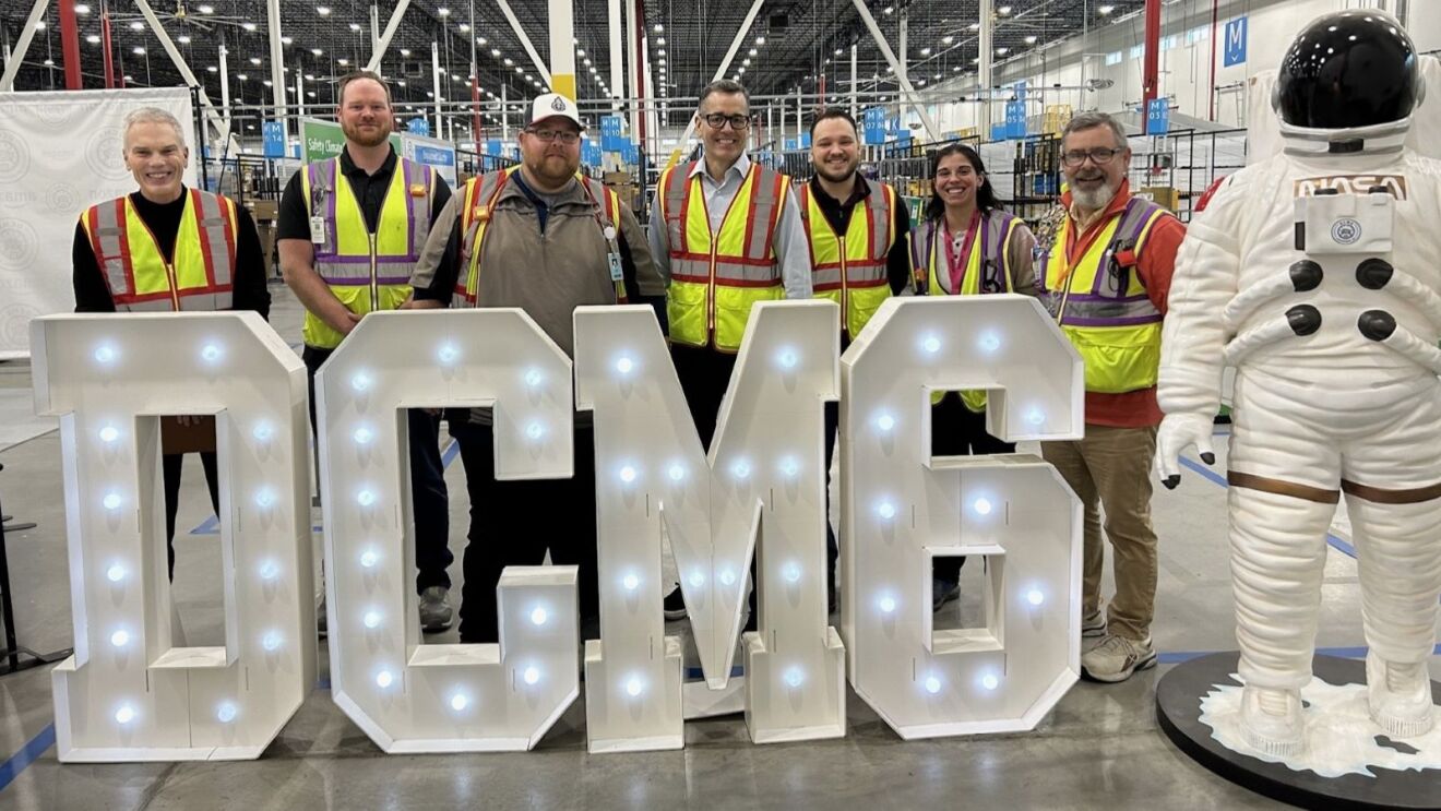 Doug Herrington visits an Amazon site posing behind illuminated 'DCM6' sign next to NASA spacesuit