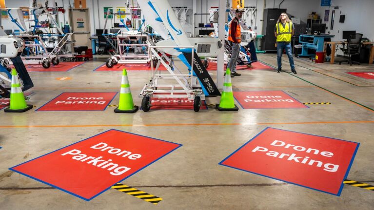 An image of multiple Amazon delivery drones parked inside the facility in Texas. There are red squares on the floor that say "drone parking."