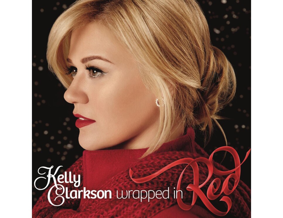 Kelly Clarkson wears her blonde hair tied behind her head, a red high-collared shirt with red scarf around her shoulders. Silver earrings, eye liner, and red lipstick completes her look. She looks to her right.