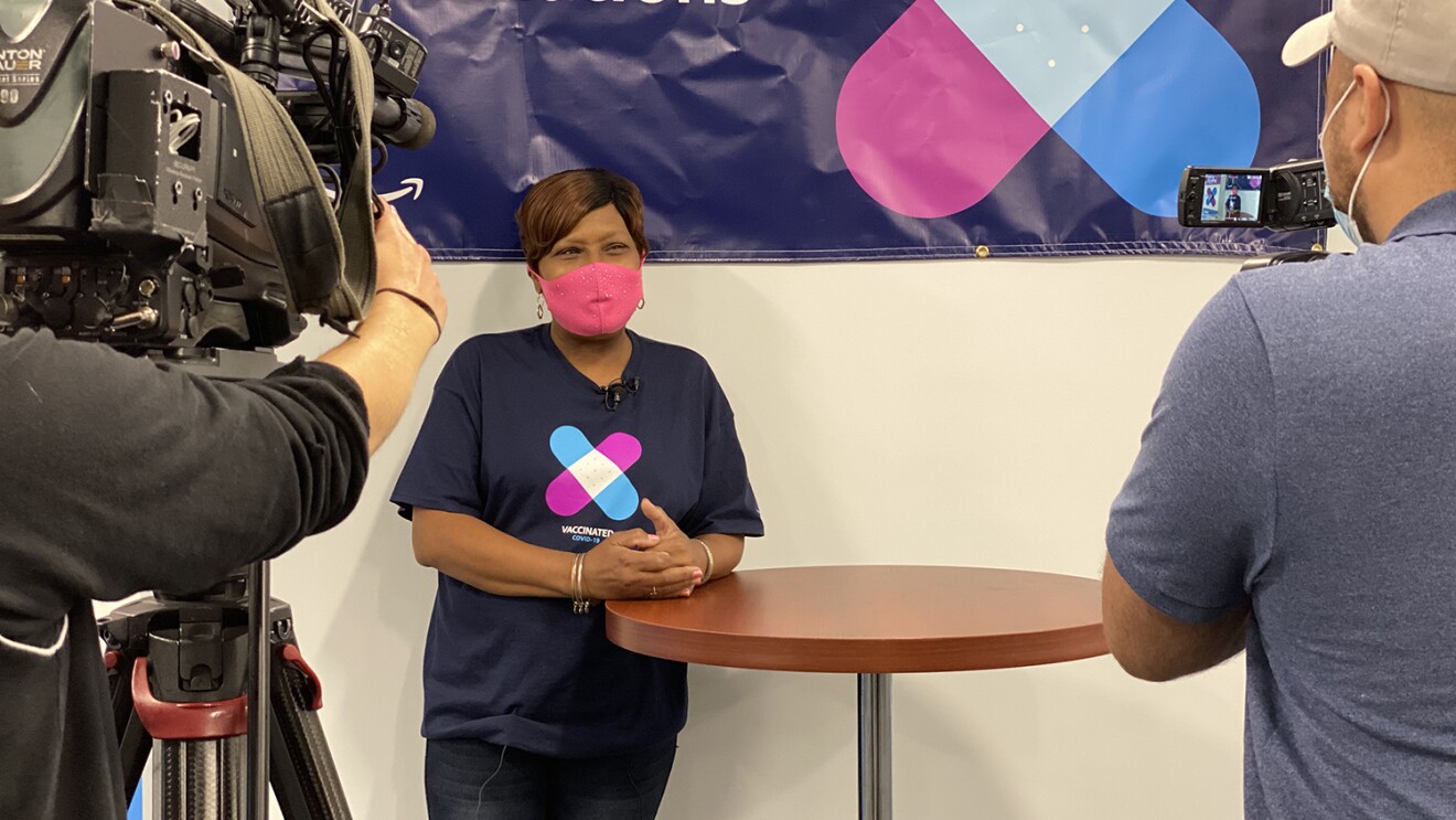An image of a woman wearing a pink mask while smiling and leaning against a tall table. She is wearing a shirt showing she has been vaccinated and being filmed by a film crew.