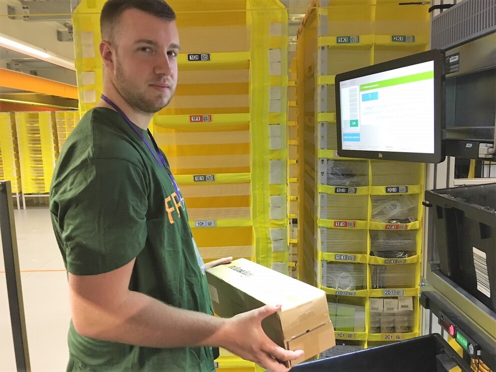 Ein Versandmitarbeiter im grünen T-Shirt ist vor einem gelben Regal zu sehen. In der Hand hält er einen Karton. Im Hintergrund ist ein Bildschirm zu sehen, der die Artikelinformationen anzeigt. Der Mann schaut freundlich-gespannt in die Kamera.