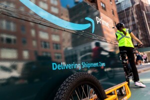 An Amazon delivery person riding a bicycle, with a bike trailer behind it. The trailer has "Delivering Shipment Zero" printed on it. 