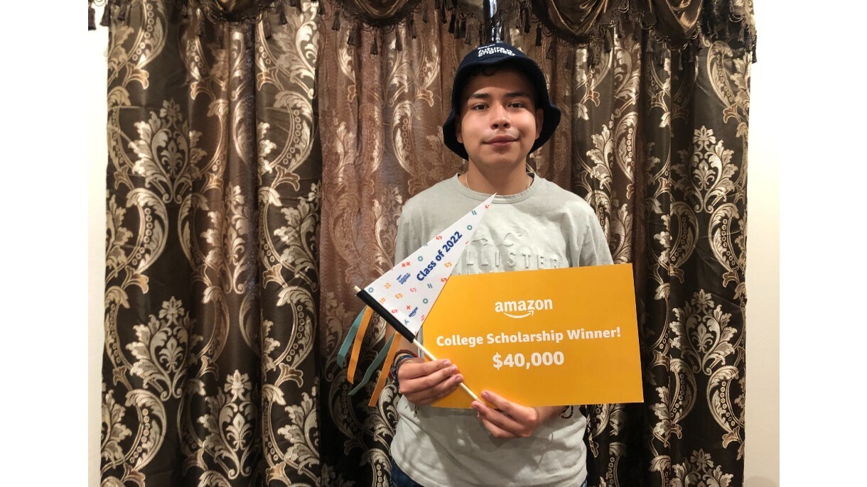 Victor Acuna stands in front of a curatin and holds up a sign that reads "College Scholarship Winner! $40,000," Acuna also holds a mini flag that reads "Class of 2022."