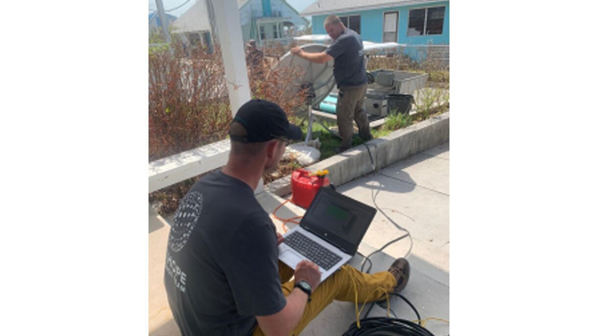 An image of a man holding up a satellite dish while another man on a laptop works on it.