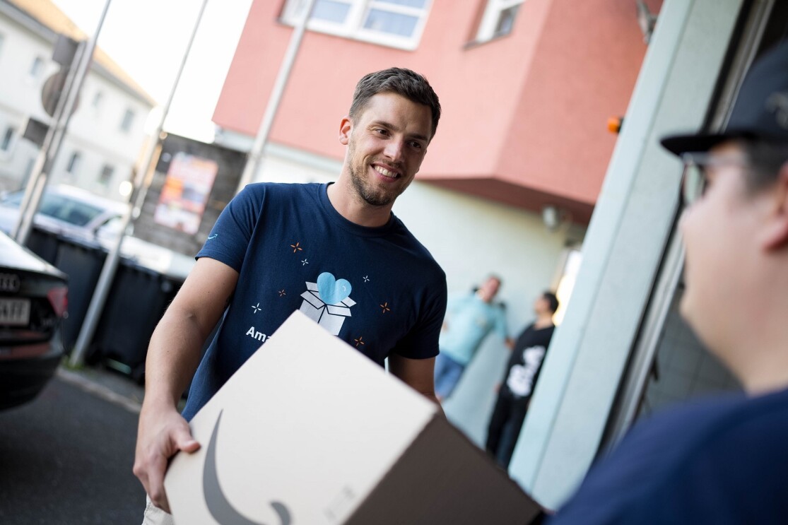 Amazon Mitarbeiter Andreas Widowitz mit Amazon Volunteers T-Shirt übergibt eine Spende im Amazon Karton an einen Kollegen der Freiwilligen Feuerwehr.